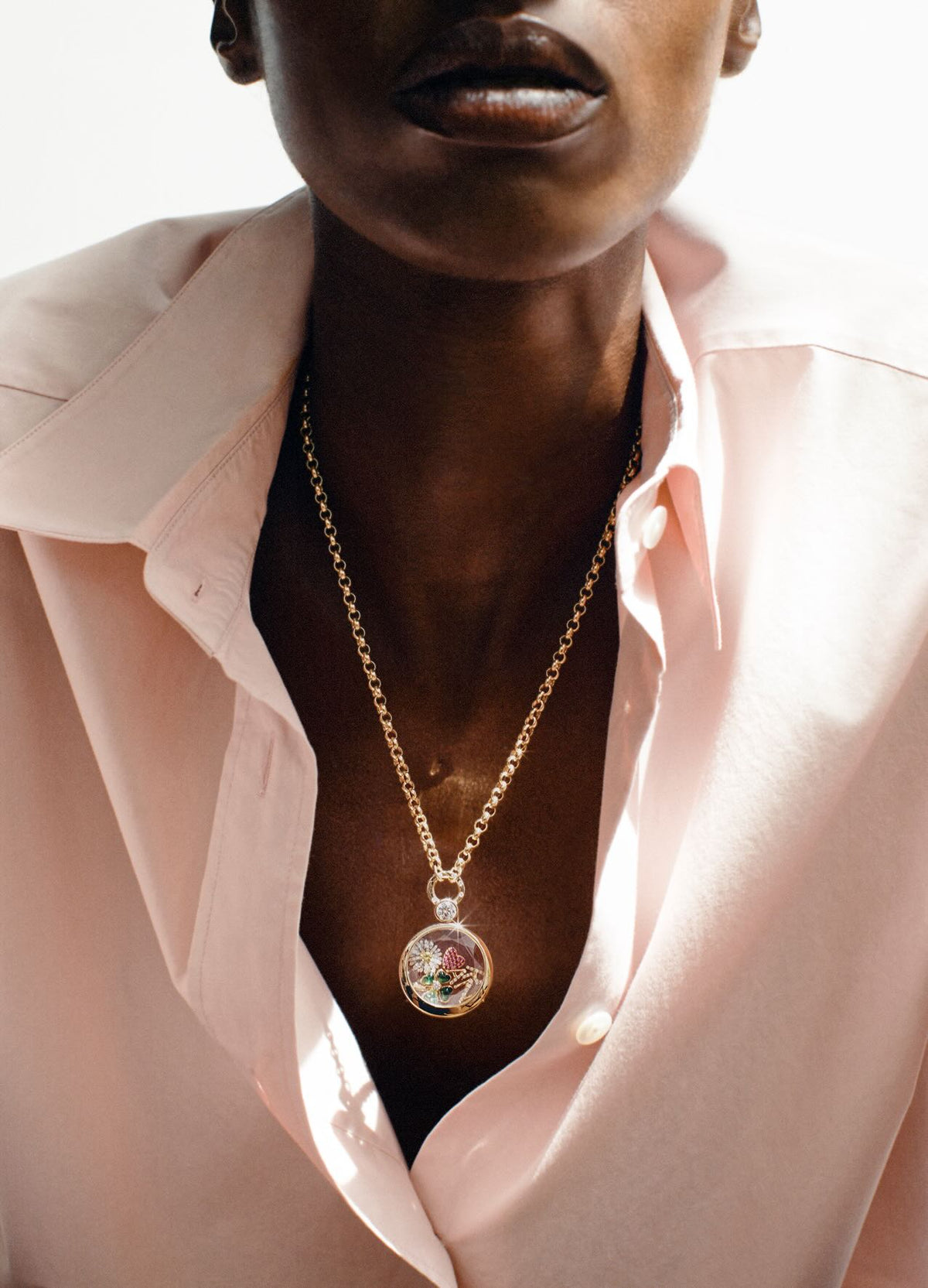 A close-up of a model wearing a gold chain necklace with a round locket pendant containing colourful charms, styled with a light pink button-down shirt.