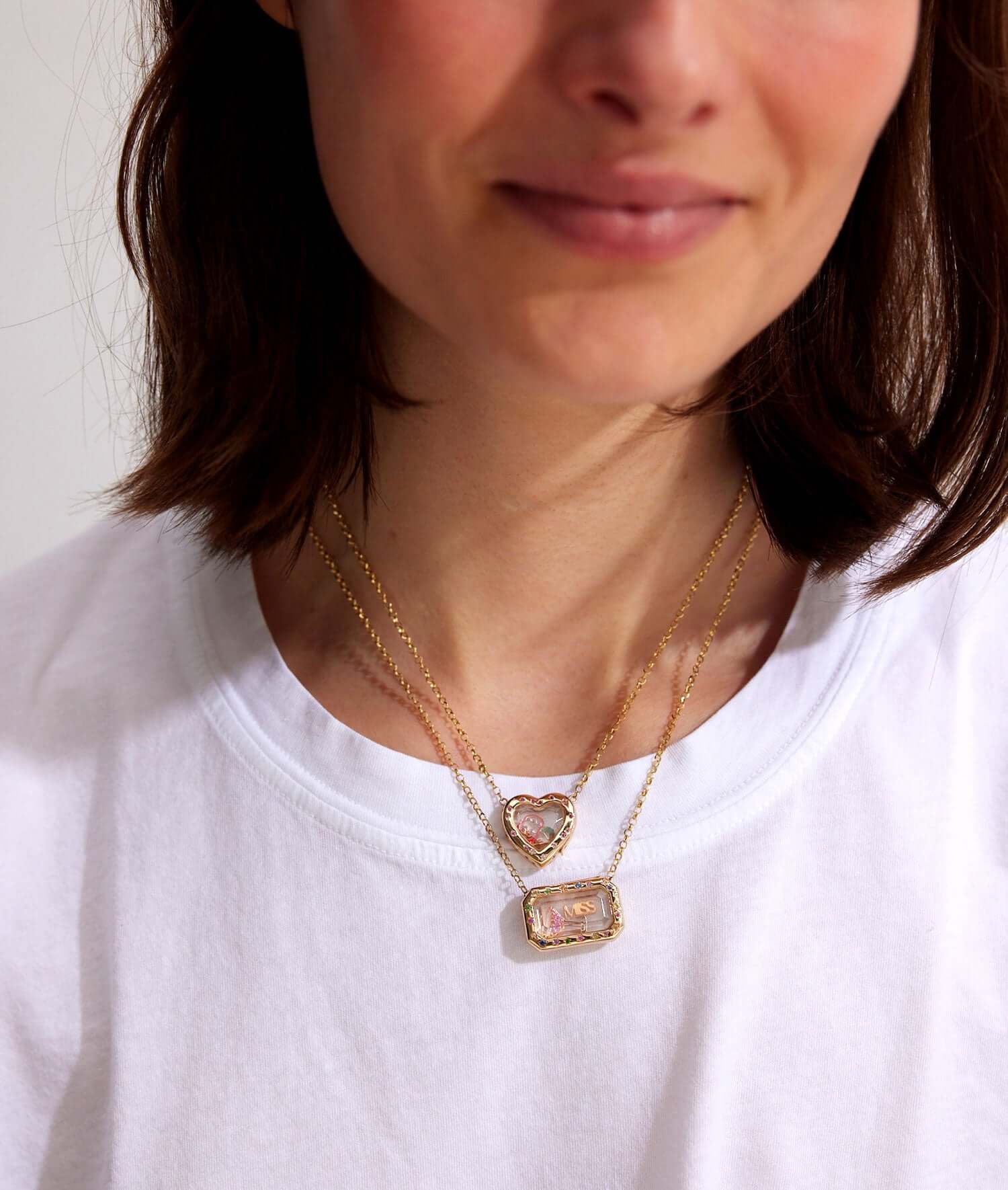 Woman wearing layered gold locket necklaces with a heart-shaped and rectangular gemstone pendant over a white shirt.