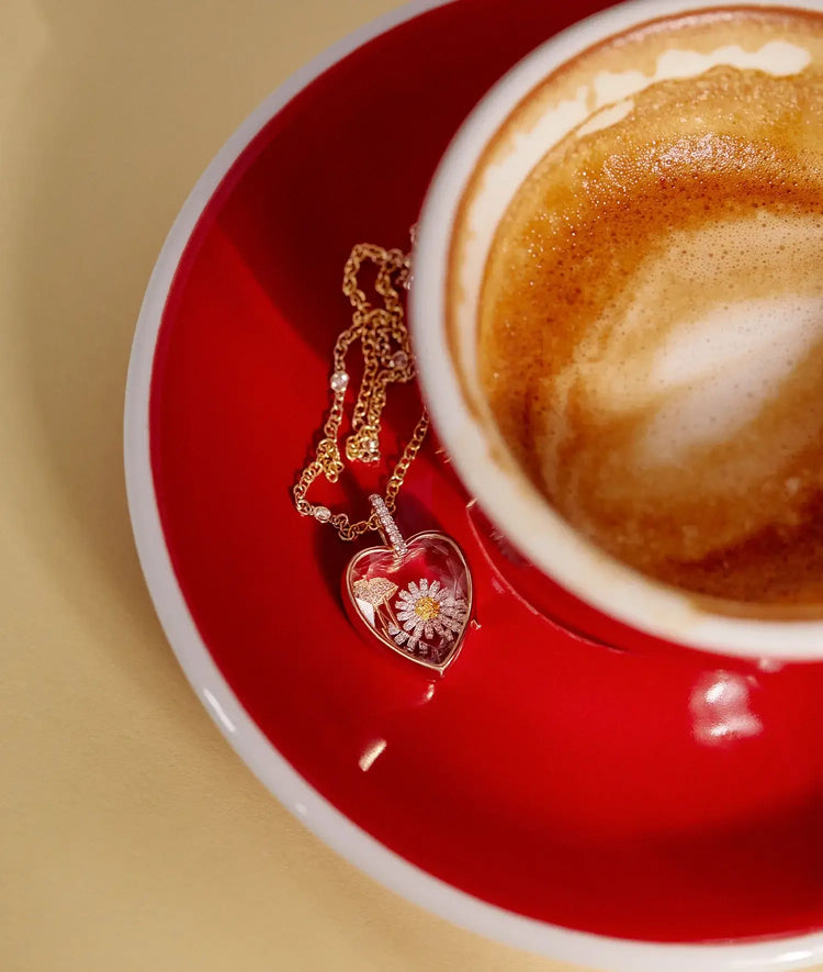 Heart-shaped charm locket necklace with flower design on a red coffee cup saucer.