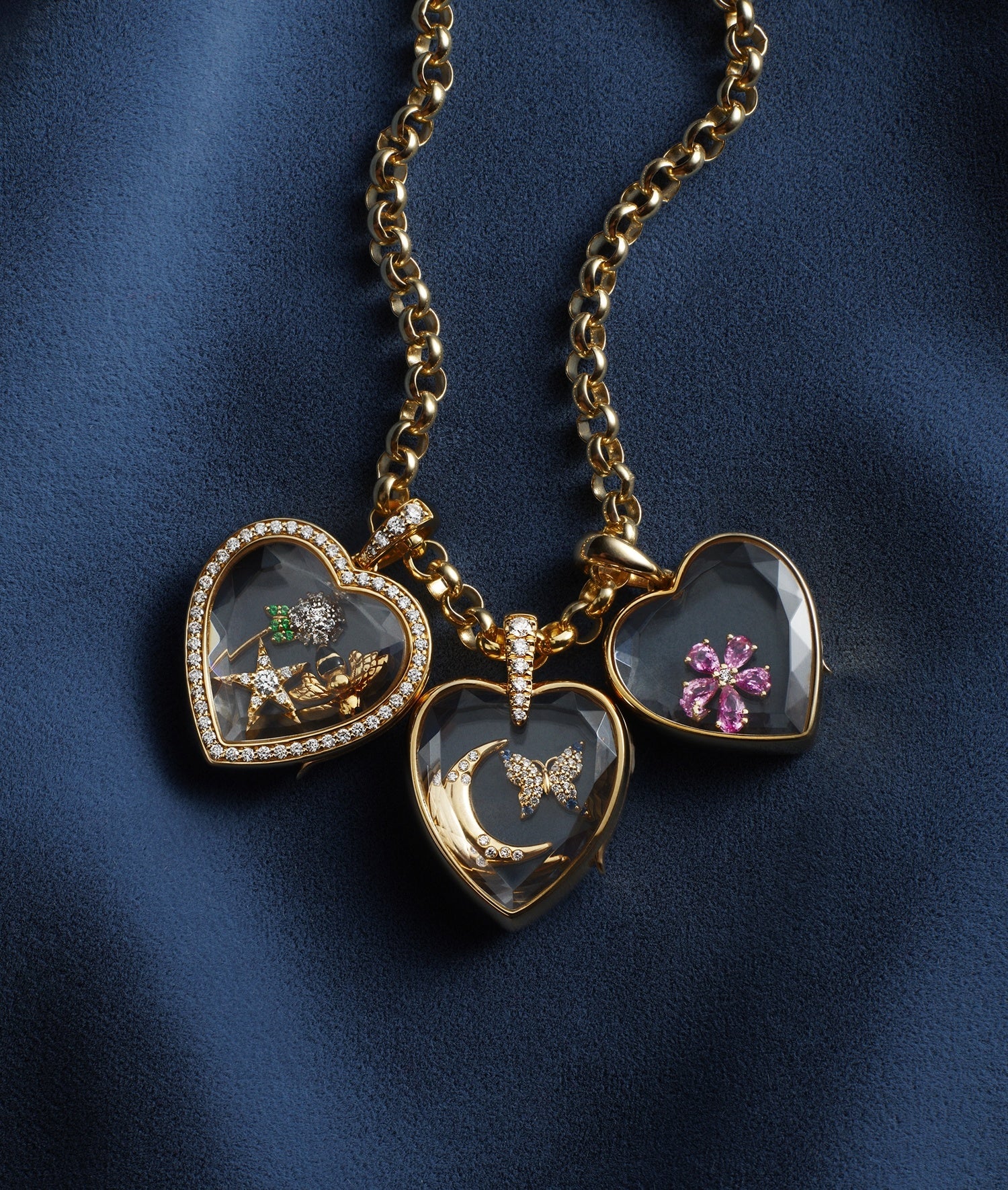 Three heart-shaped gold locket pendants with clear crystal facade, hanging from a gold chain on a dark blue fabric background. One locket shows a dandelion, one shows a pink flower, and one shows a gold crescent moon and butterfly.