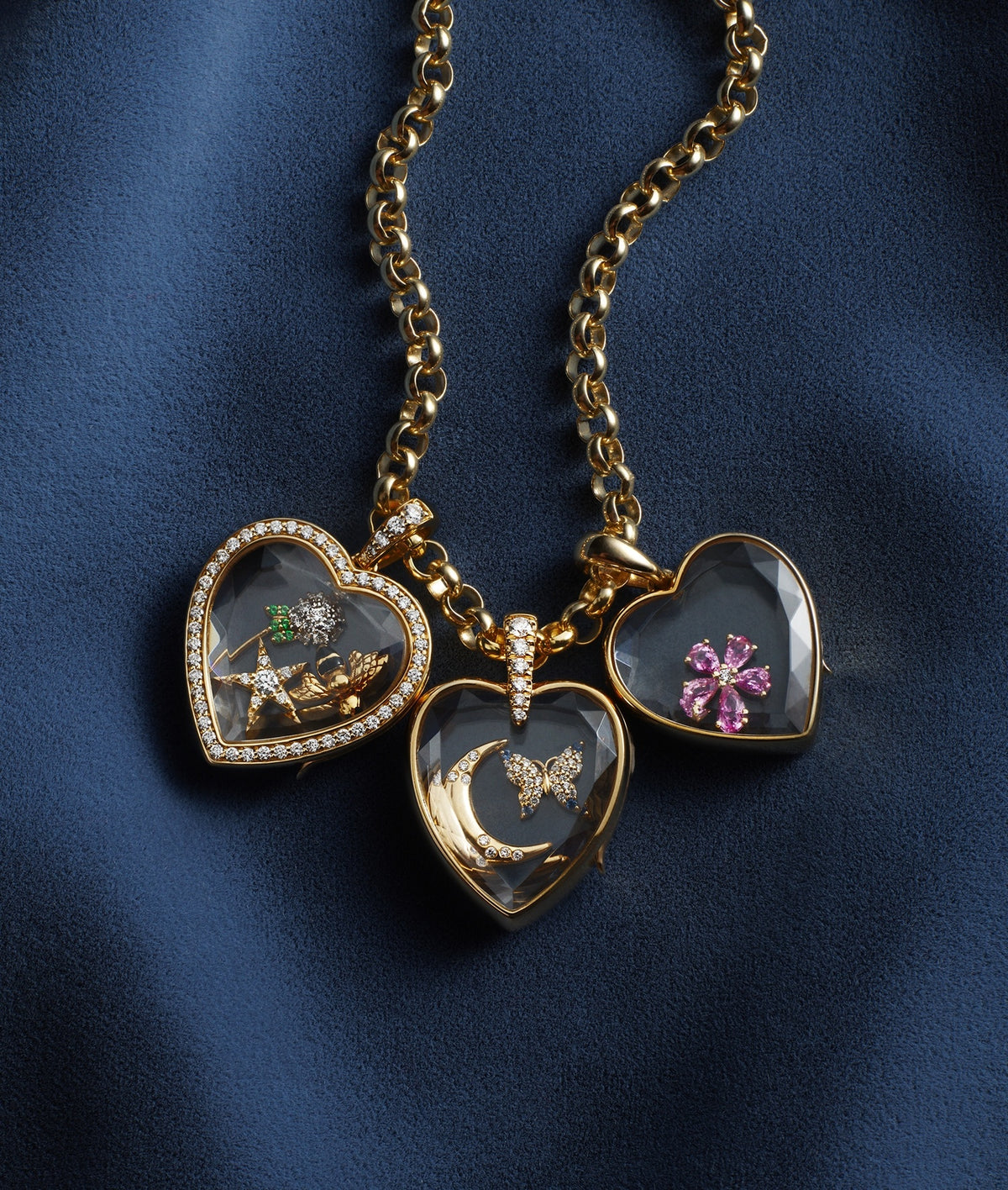 Three heart-shaped gold locket pendants with clear crystal facade, hanging from a gold chain on a dark blue fabric background. One locket shows a dandelion, one shows a pink flower, and one shows a gold crescent moon and butterfly.