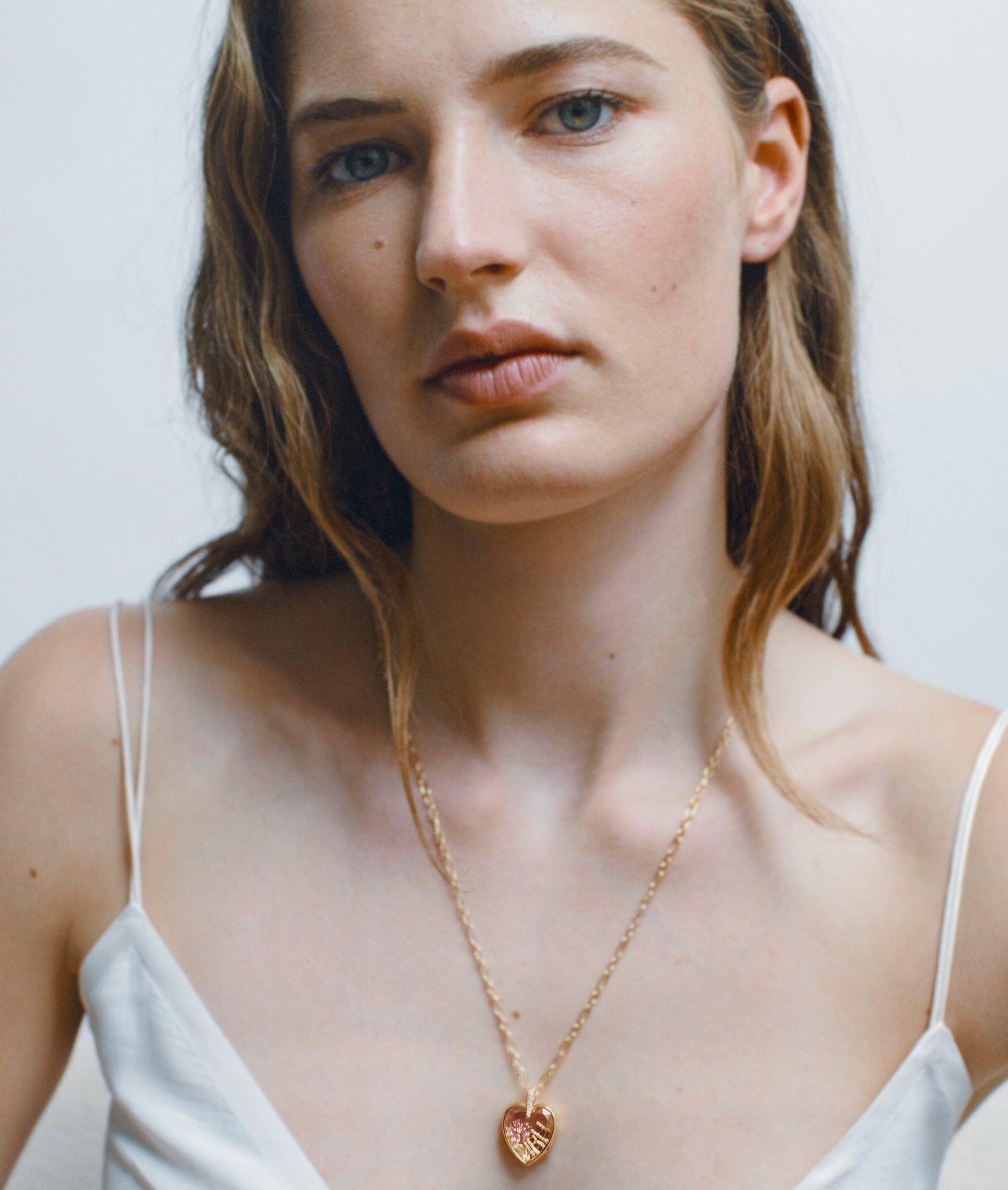 A close-up of a woman wearing a delicate gold chain necklace with a small heart-shaped locket Amate pendant, styled with a white spaghetti-strap top against a light grey background.