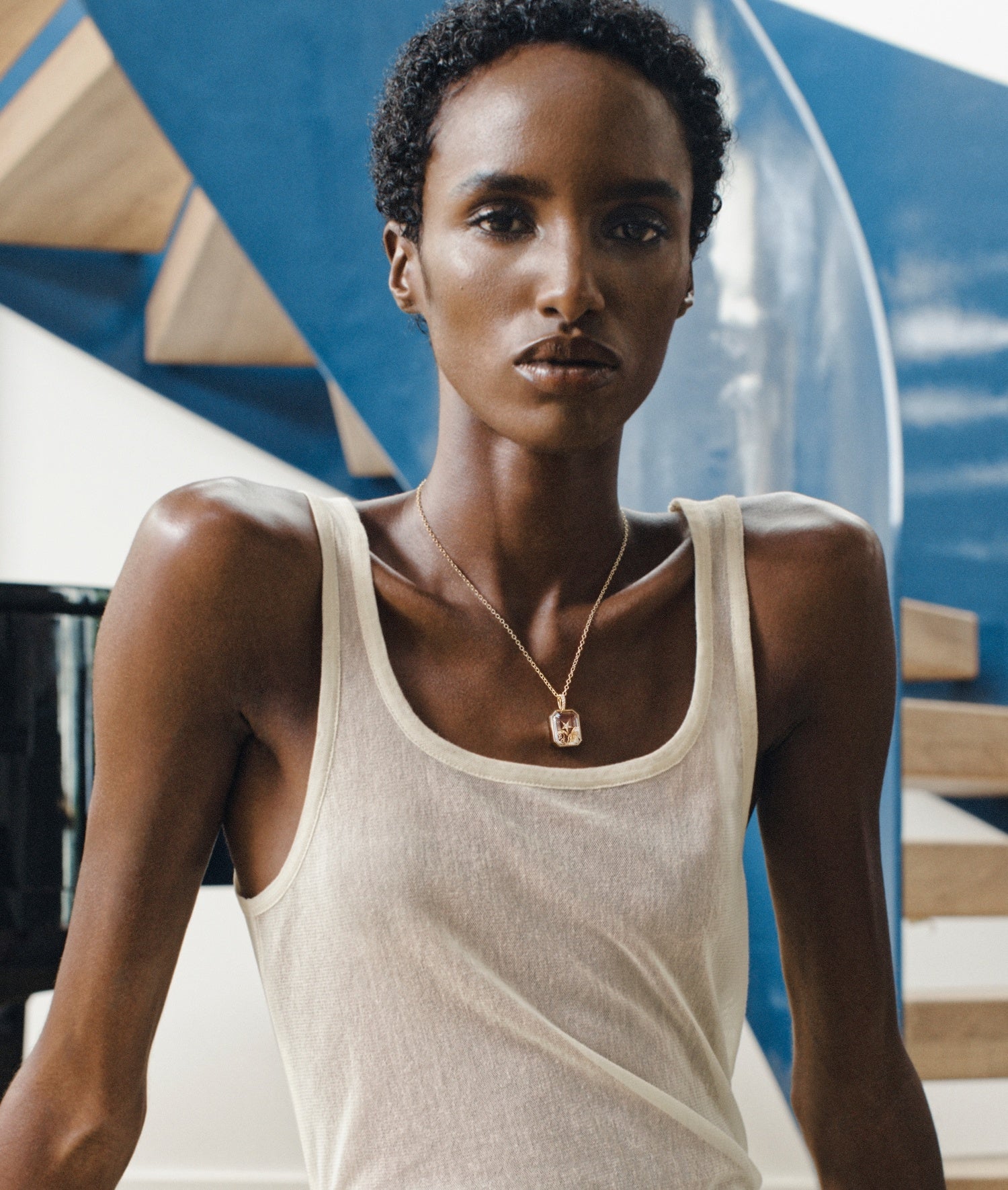 A model wearing a cream-colored ribbed tank top and a yellow gold locket pendant necklace with a clear faceted crystal facade, posed in front of a modern blue and white architectural background.