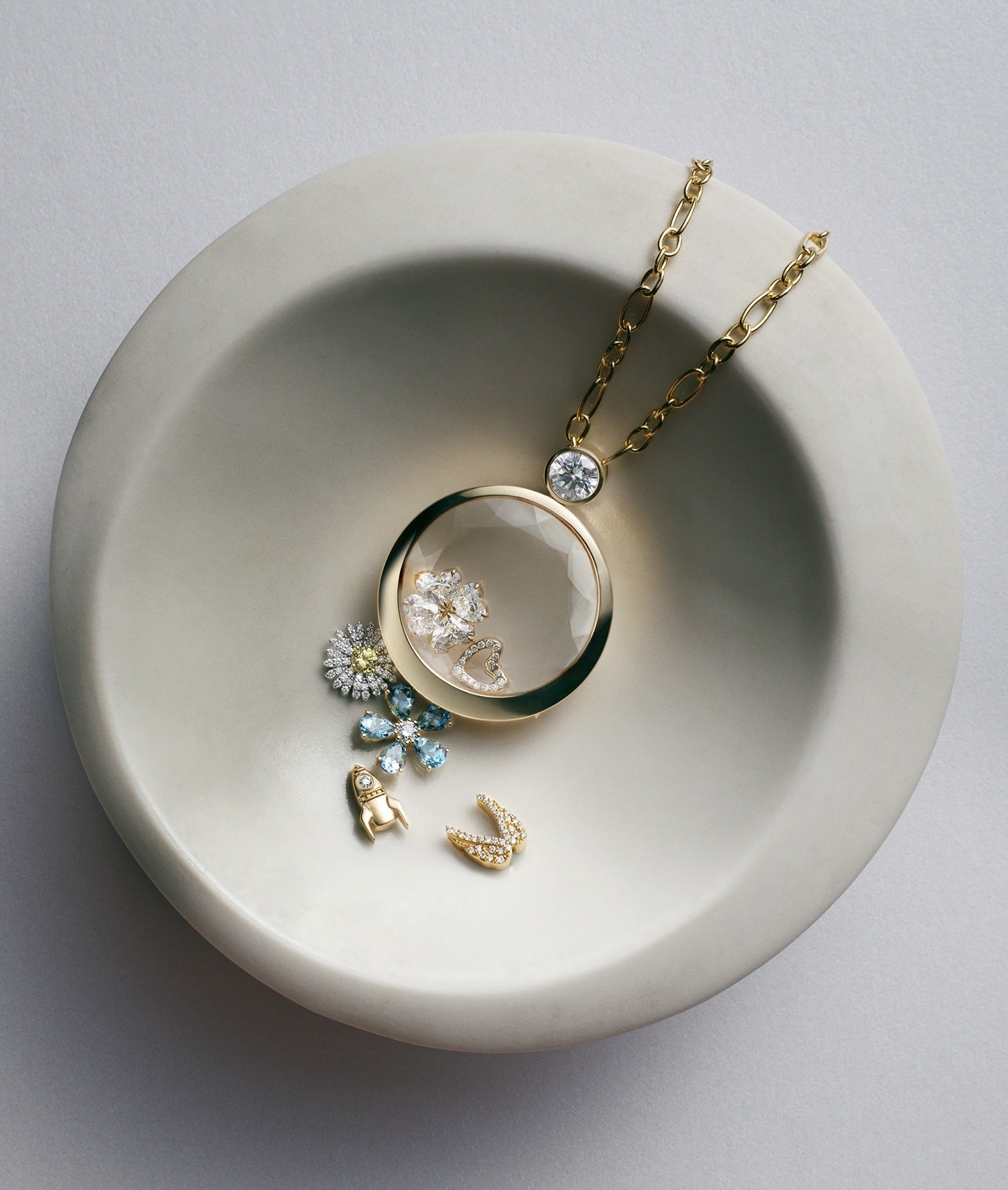 A gold ound locket necklace with a clear crystal facade containing diamond flower petals, displayed on a light grey ceramic dish with small matching gold charms beside it.