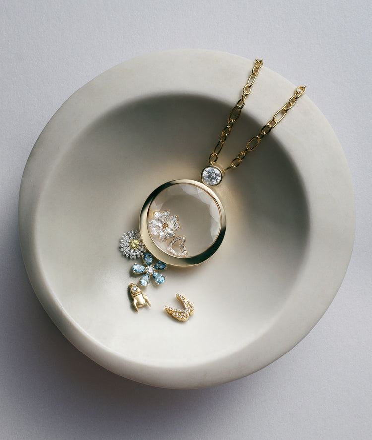 A gold ound locket necklace with a clear crystal facade containing diamond flower petals, displayed on a light grey ceramic dish with small matching gold charms beside it.