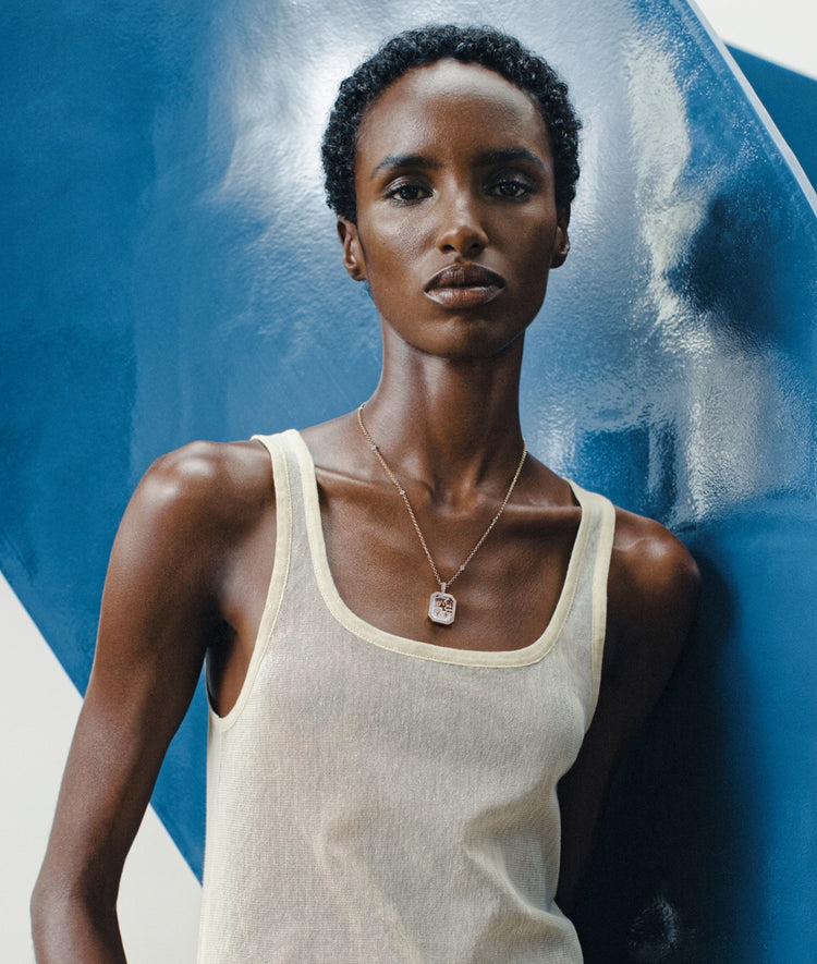 A model wearing a cream-colored ribbed tank top and a loquet gold pendant necklace with a clear rectangular crystal facade, posed against a textured blue and white background.