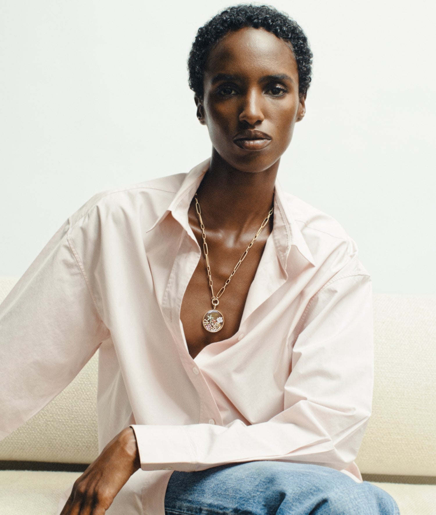 A model wearing a pale pink button-down shirt and blue jeans, accessorized with a gold clip chain featuring a round jupiter locket pendant, seated against a light beige wall.