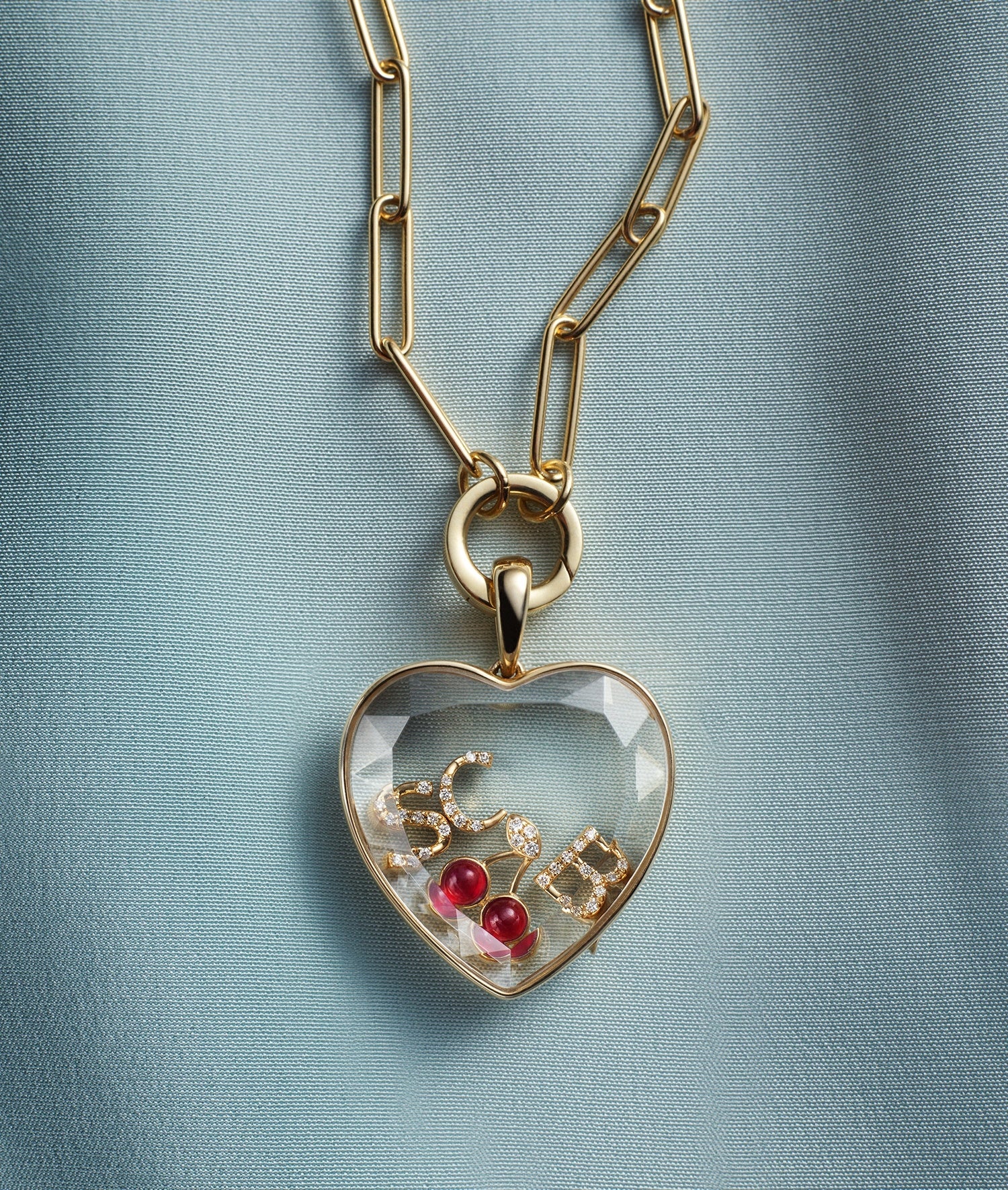 Large heart locket pendant styled with a clip chain in yellow gold and a round clip with initial and cherry charms on a textured background