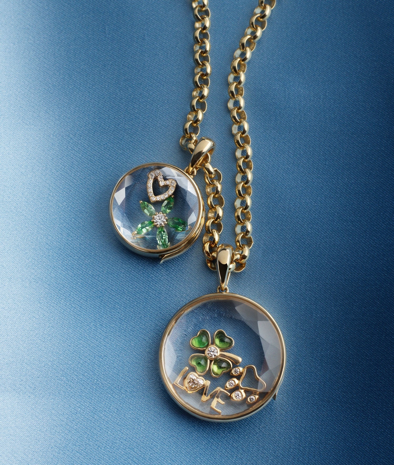 Large round and medium round locket pendants styled with diamond and emerald charms with a yellow gold chain on a blue textured background