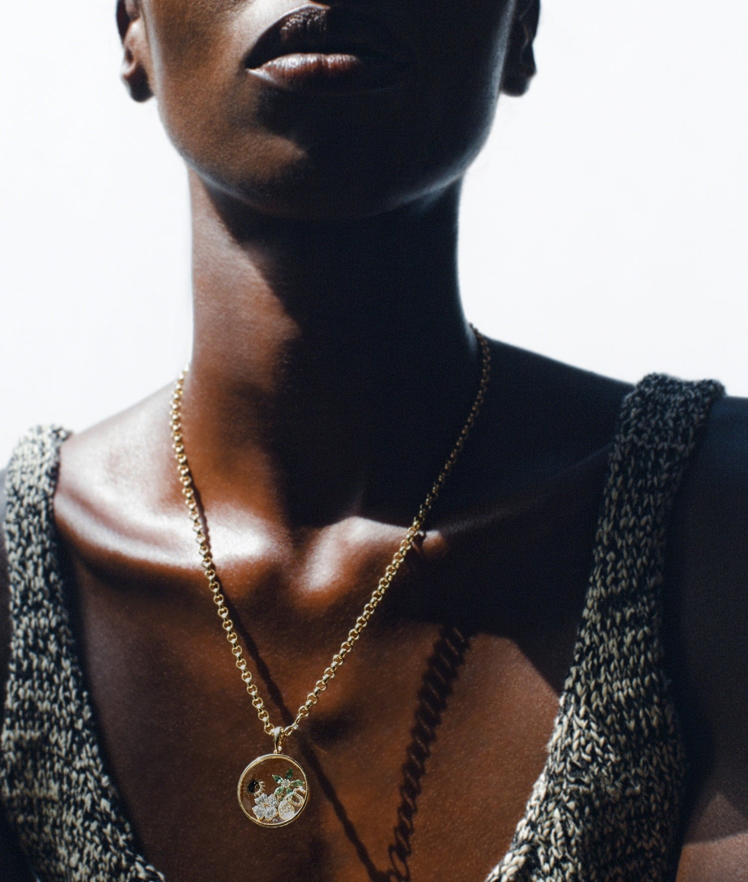 A close-up lifestyle shot of a model wearing a gold chain necklace with a large round locket pendant styled with diamond charms against a high-contrast bright white background.