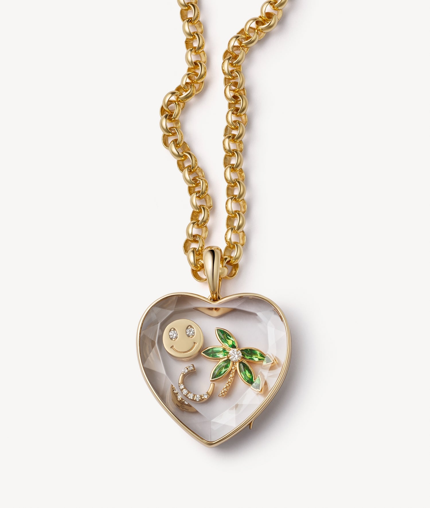A gold heart-shaped locket necklace with a faceted clear crystal facade containing a gold smiley face charm, a green palm tree charm, and a gold C letter with diamond stones, shown on a light grey background.