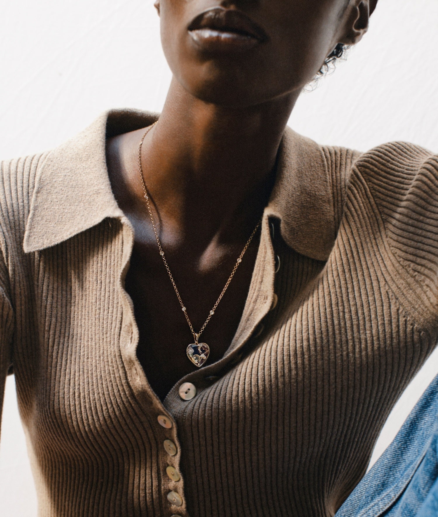 A close-up lifestyle shot of a model wearing a delicate gold chain necklace with a heart-shaped locket pendant featuring diamond charms styled over a brown ribbed knit polo shirt.