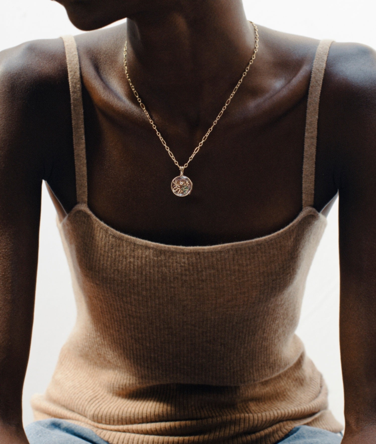 A close-up of a model wearing a delicate gold chain necklace with a round locket pendant, styled with a beige ribbed knit camisole top.