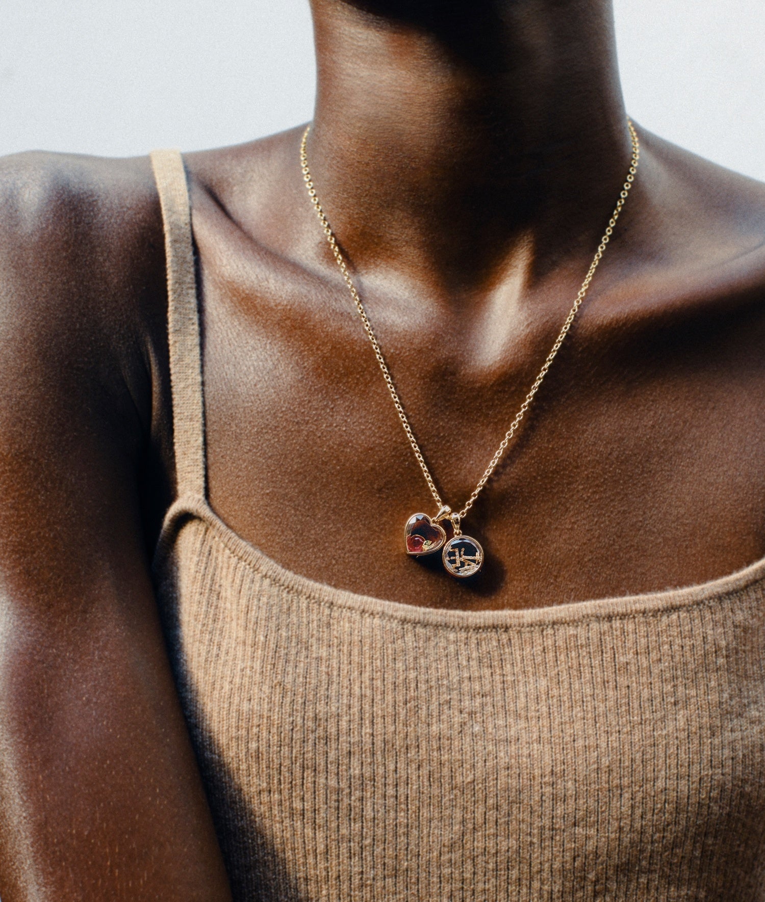 Mini lockets pendant heart and round shaped on a yellow gold chain worn my a model with a beige tank top
