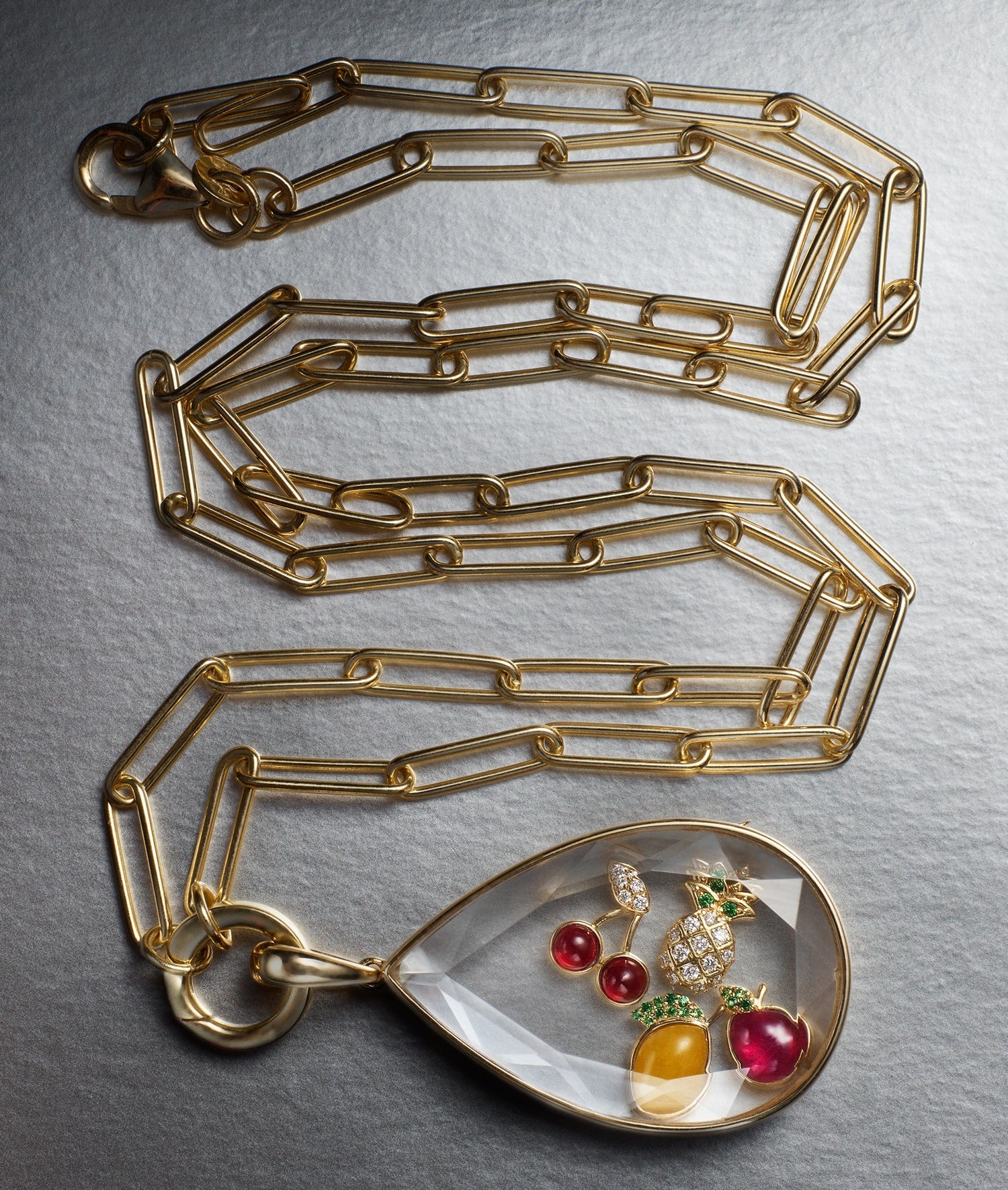 A gold chain necklace with a teardrop-shaped crystal locket pendant containing colorful fruit and berry charms, displayed on a textured gray background.
