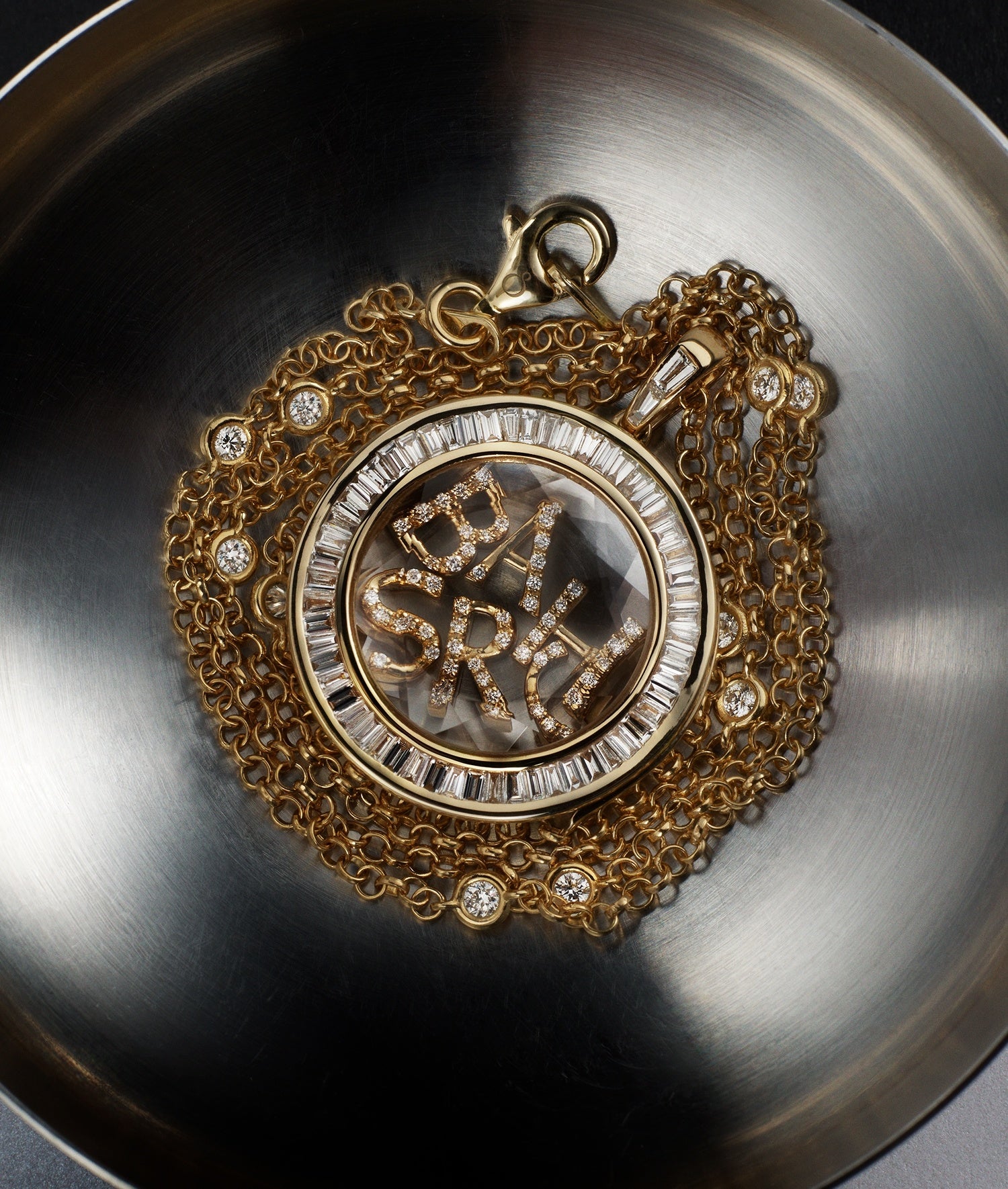 Orb Baguette Diamond locket pendant styled with initial diamond charms and a diamond chain in yellow gold on a aluminium bowl