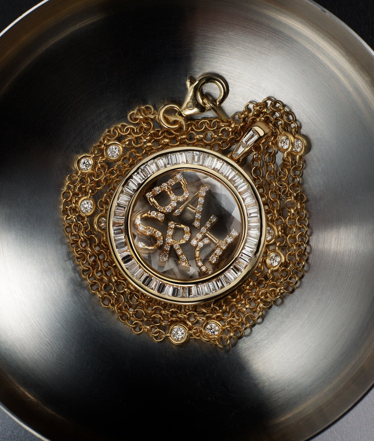 Orb Baguette Diamond locket pendant styled with initial diamond charms and a diamond chain in yellow gold on a aluminium bowl