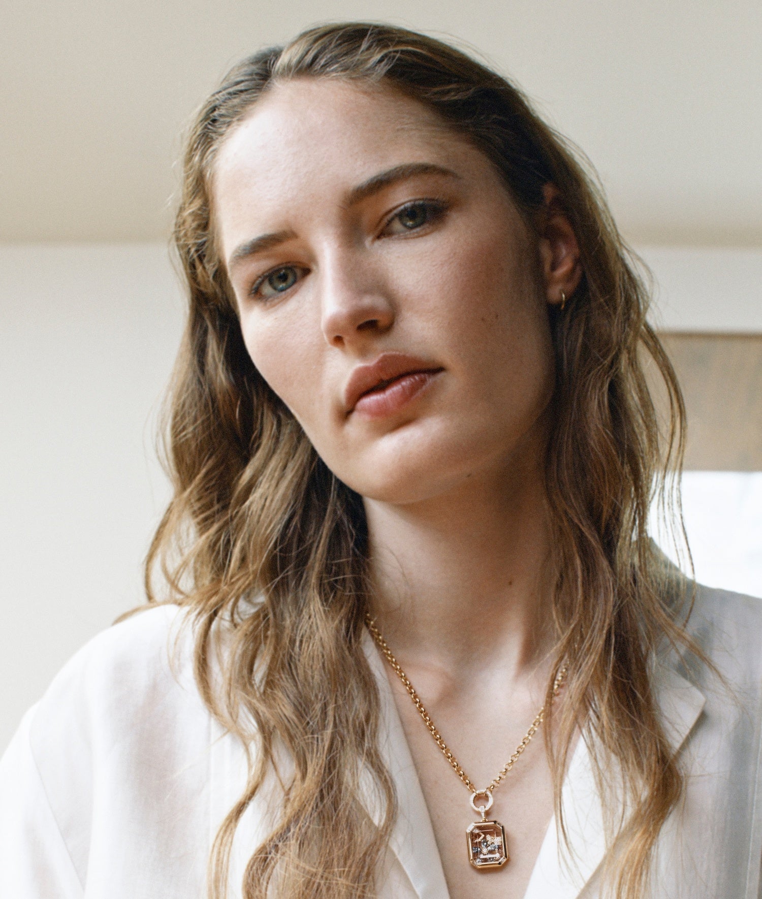 A close-up portrait of a woman with long wavy hair wearing a delicate gold chain with a baguette quattro locket by loquet london, against a soft off-white background.