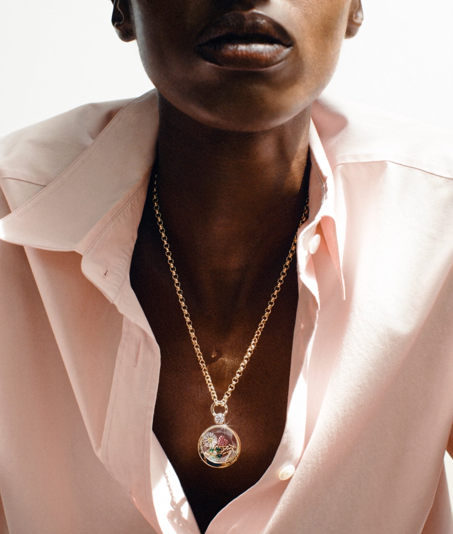 A close-up lifestyle shot of a model wearing a gold chain necklace with a solitaire diamond locket pendant containing colorful flower charms, styled with a light pink button-down shirt