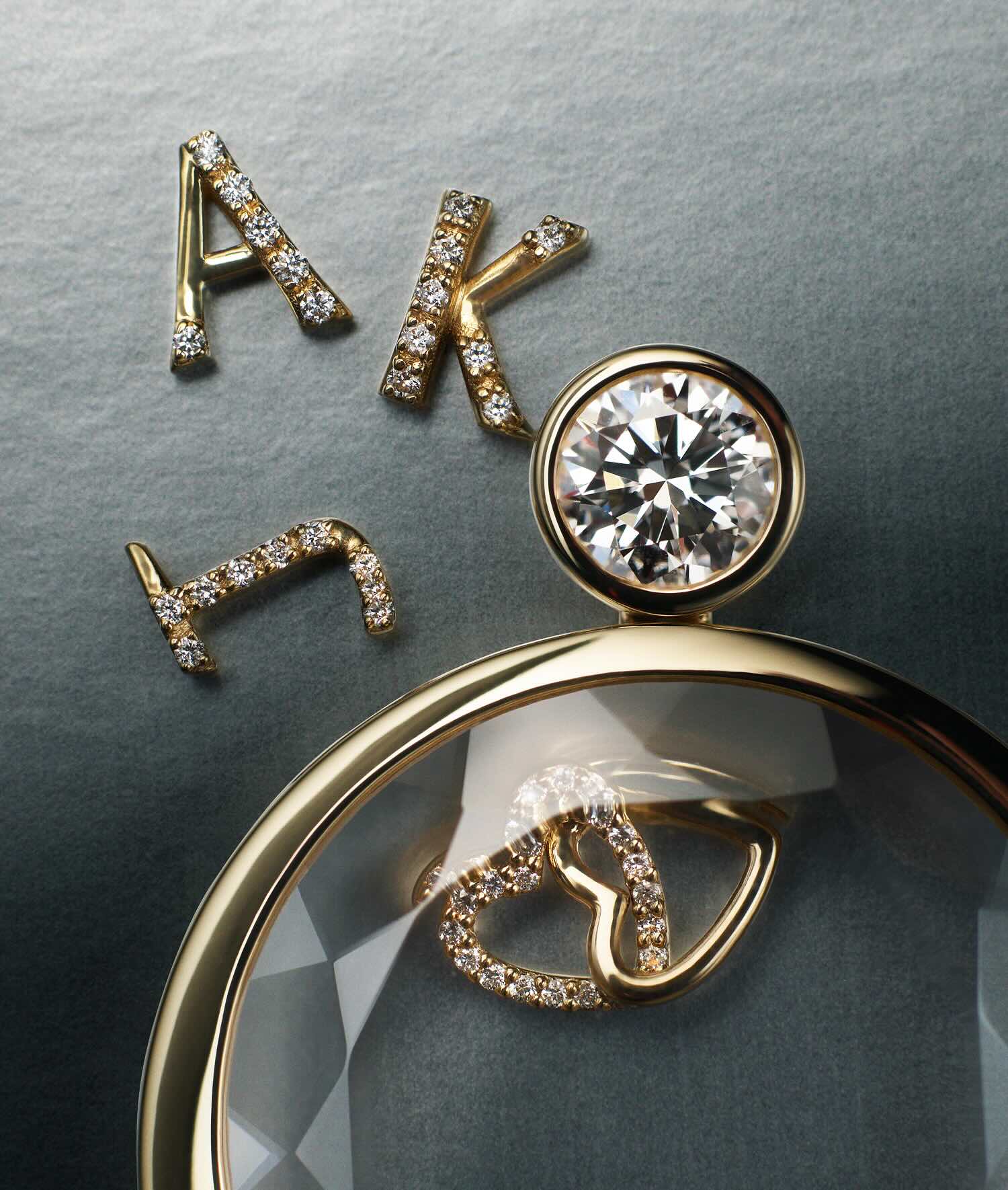 Gold letter initial charms set with diamonds, displayed alongside a round solitaire locket pendant with a interlinked hearts charm on a textured grey background