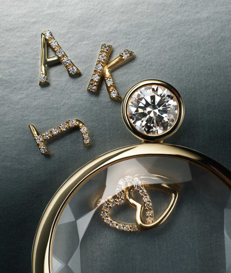 Gold letter initial charms set with diamonds, displayed alongside a round solitaire locket pendant with a interlinked hearts charm on a textured grey background