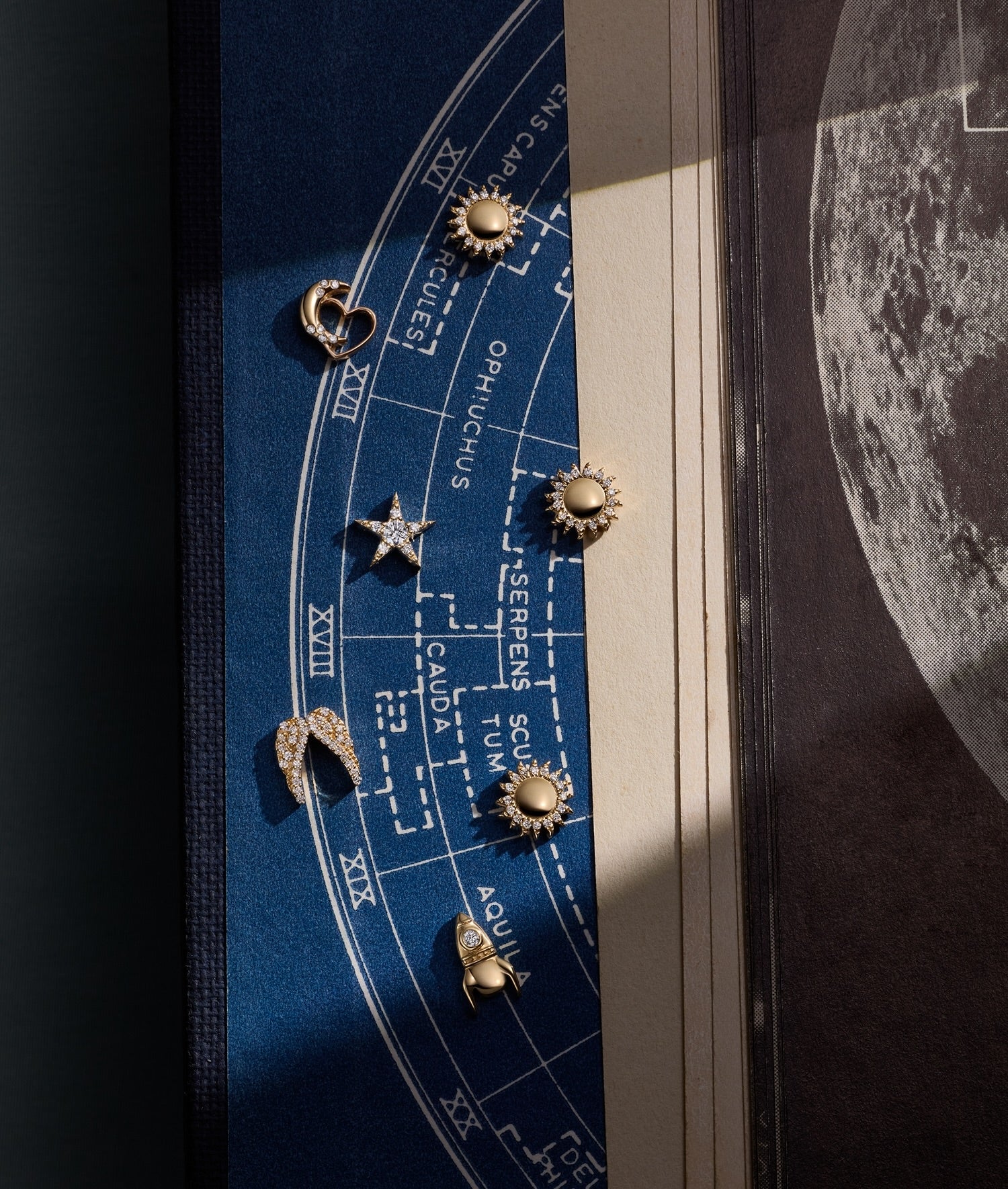 A close-up of several gold charms arranged on a blue zodiac chart print, with a book or magazine page and moon imagery visible in the background.