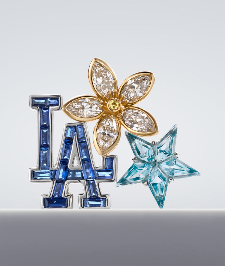 A luxury piece featuring a gold flower charm with marquise-cut diamonds, a blue 'LA' letter charm set with blue sapphires, and a light blue faceted star charm, all on a neutral grey background.