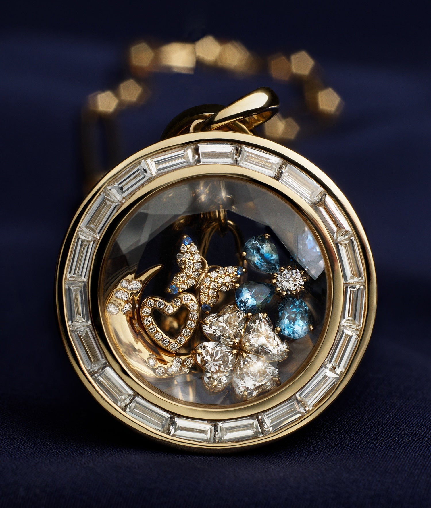 A round gold locket pendant necklace with a diamond baguette border, containing a mix of gold and blue gemstones charms, resting on a dark navy blue fabric background.