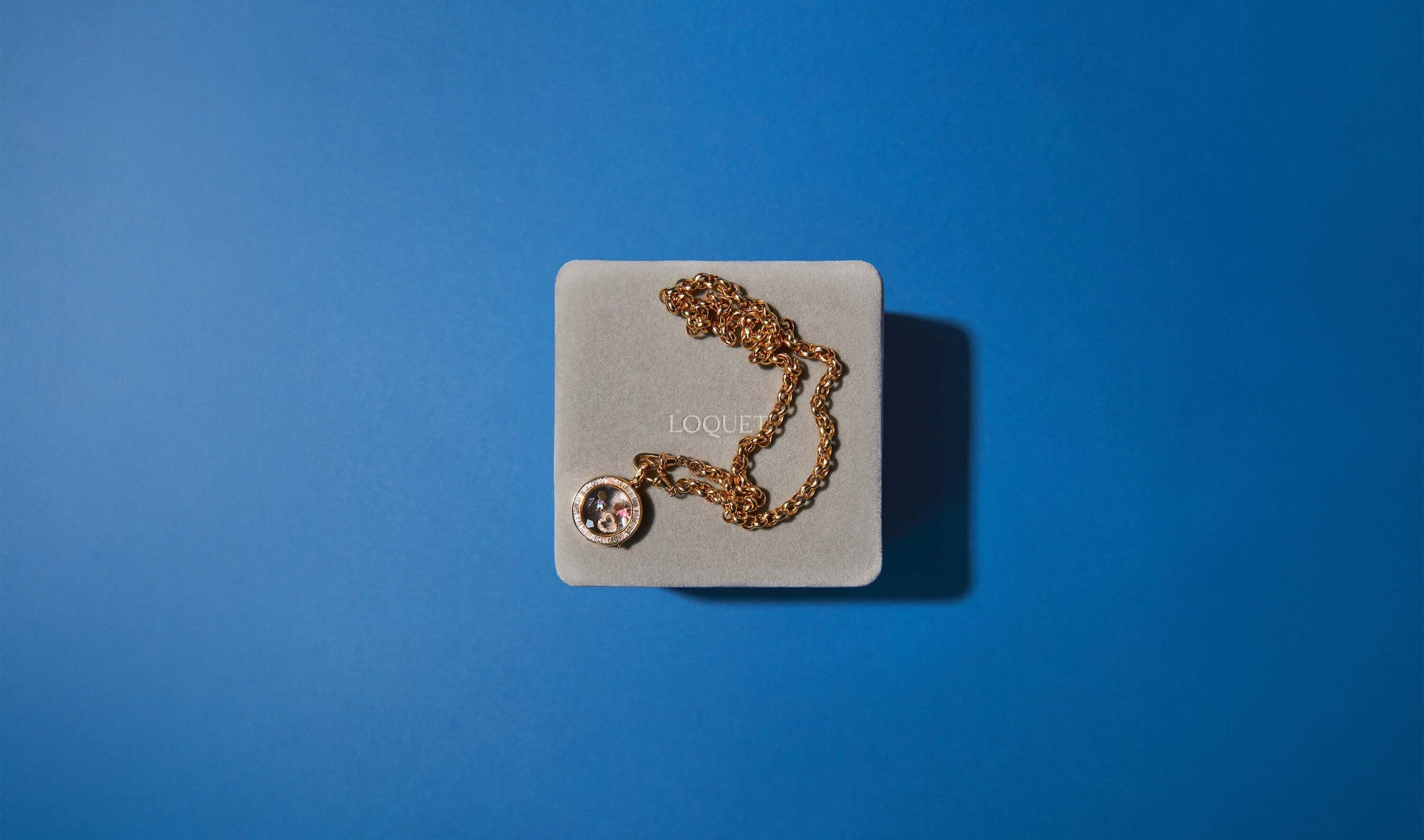 Grey velvet iconic Loquet London gift box with a baguette orb diamond locket on a chain on a electric blue background