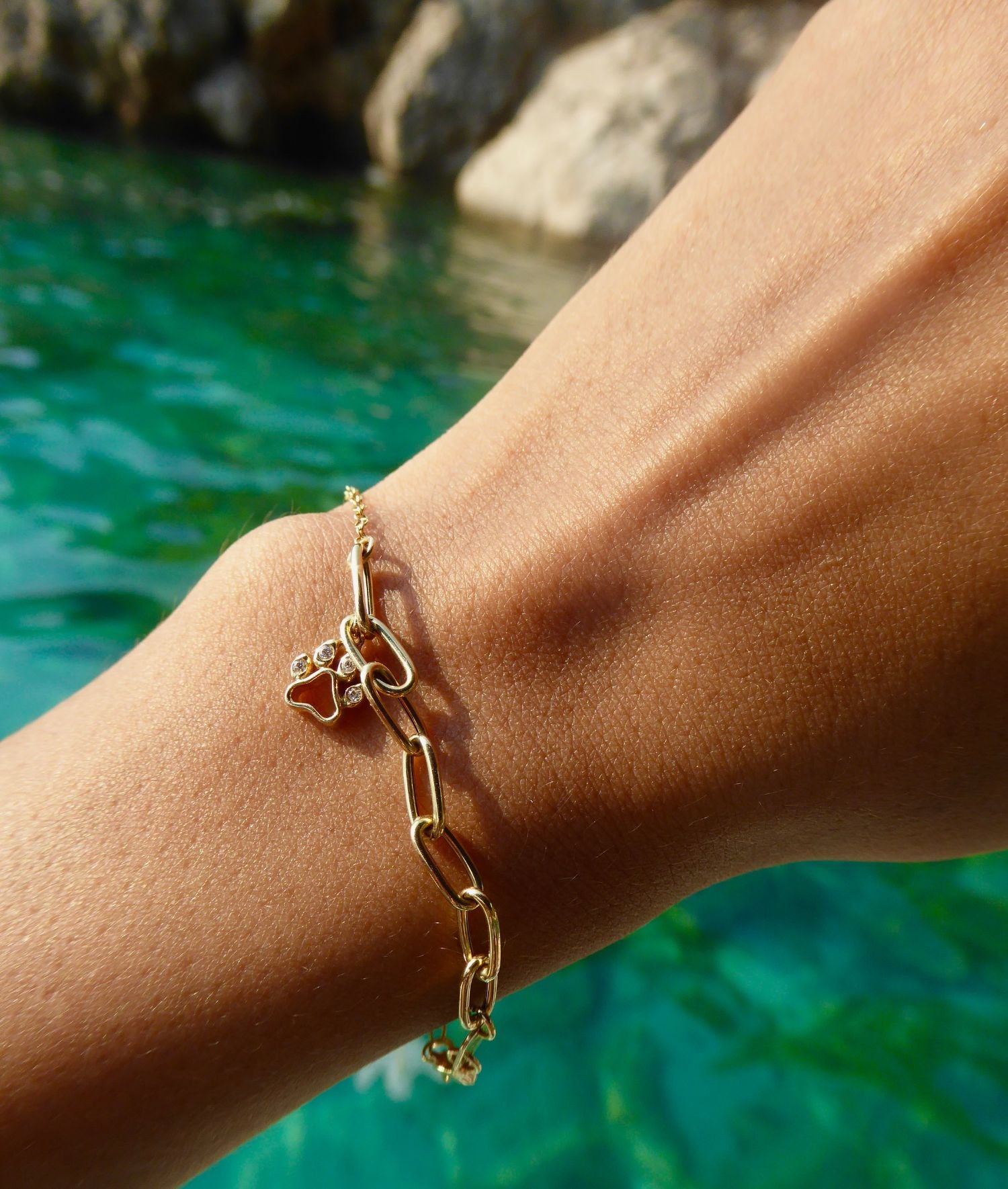 Gold chain bracelet with heart charm on wrist by clear turquoise water.