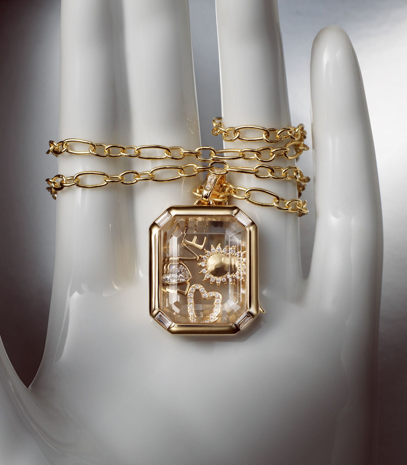 A gold chain with a rectangular locket pendant containing miniature celestial and floral diamond charms, displayed on a white mannequin hand with a gold chain draped around it.