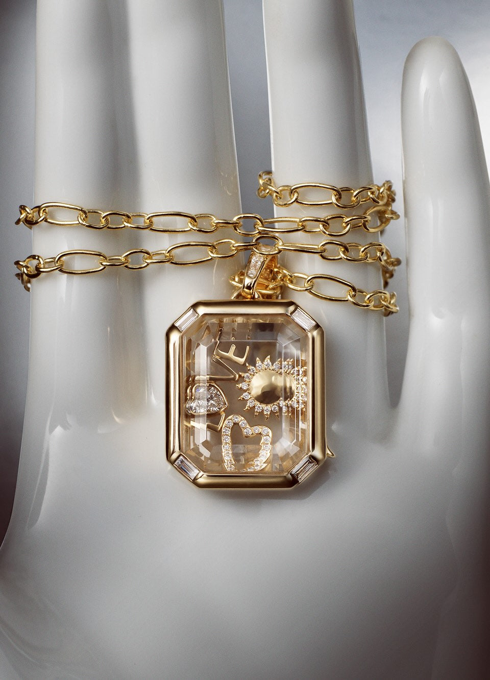 A gold chain with a rectangular locket pendant containing miniature celestial and floral diamond charms, displayed on a white mannequin hand with a gold chain draped around it.