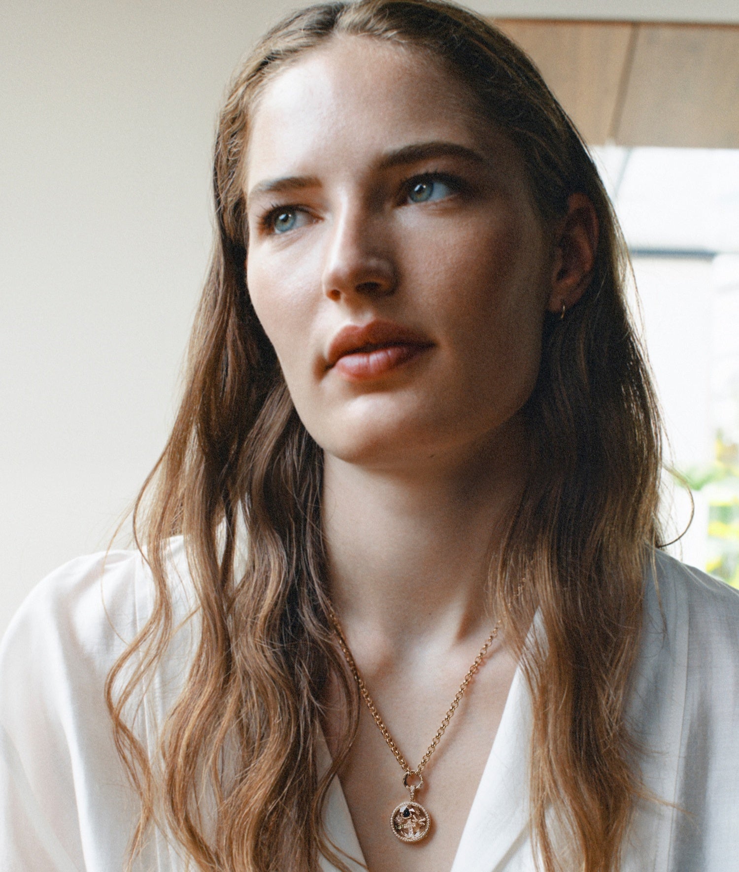 Portrait of a model wearing a diamond round locket pendant on a chain styled with charms with a white top shirt