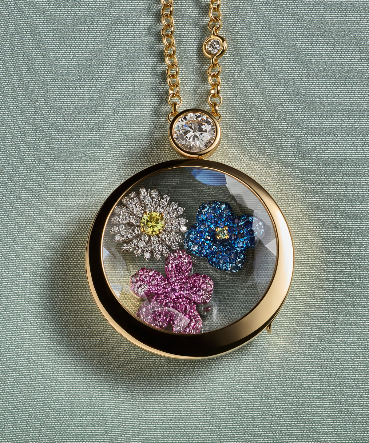 Gold necklace with a round locket pendant featuring colourful floral charms on a light blue background