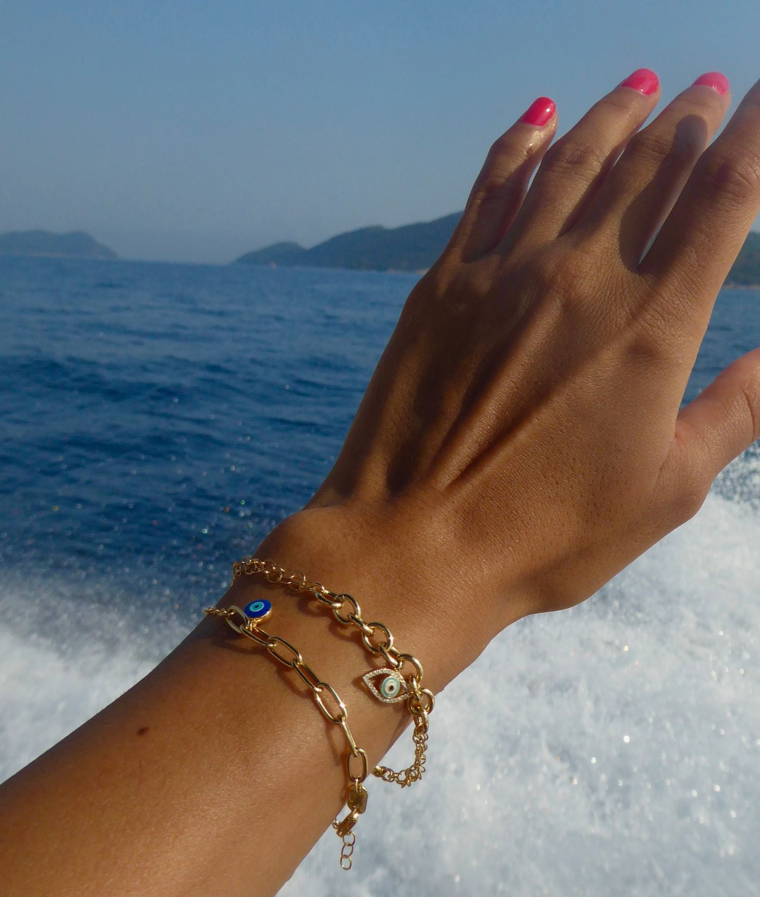 Hand with gold bracelets featuring evil eye charms held against ocean background.